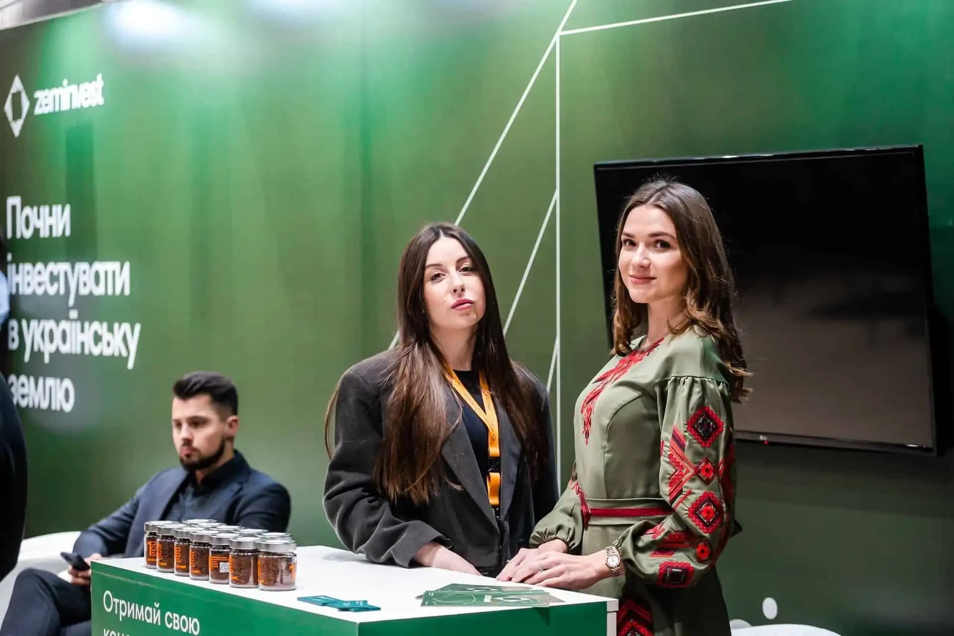 Two women standing at a booth with a green "zeminvest" backdrop, showcasing products on a counter.