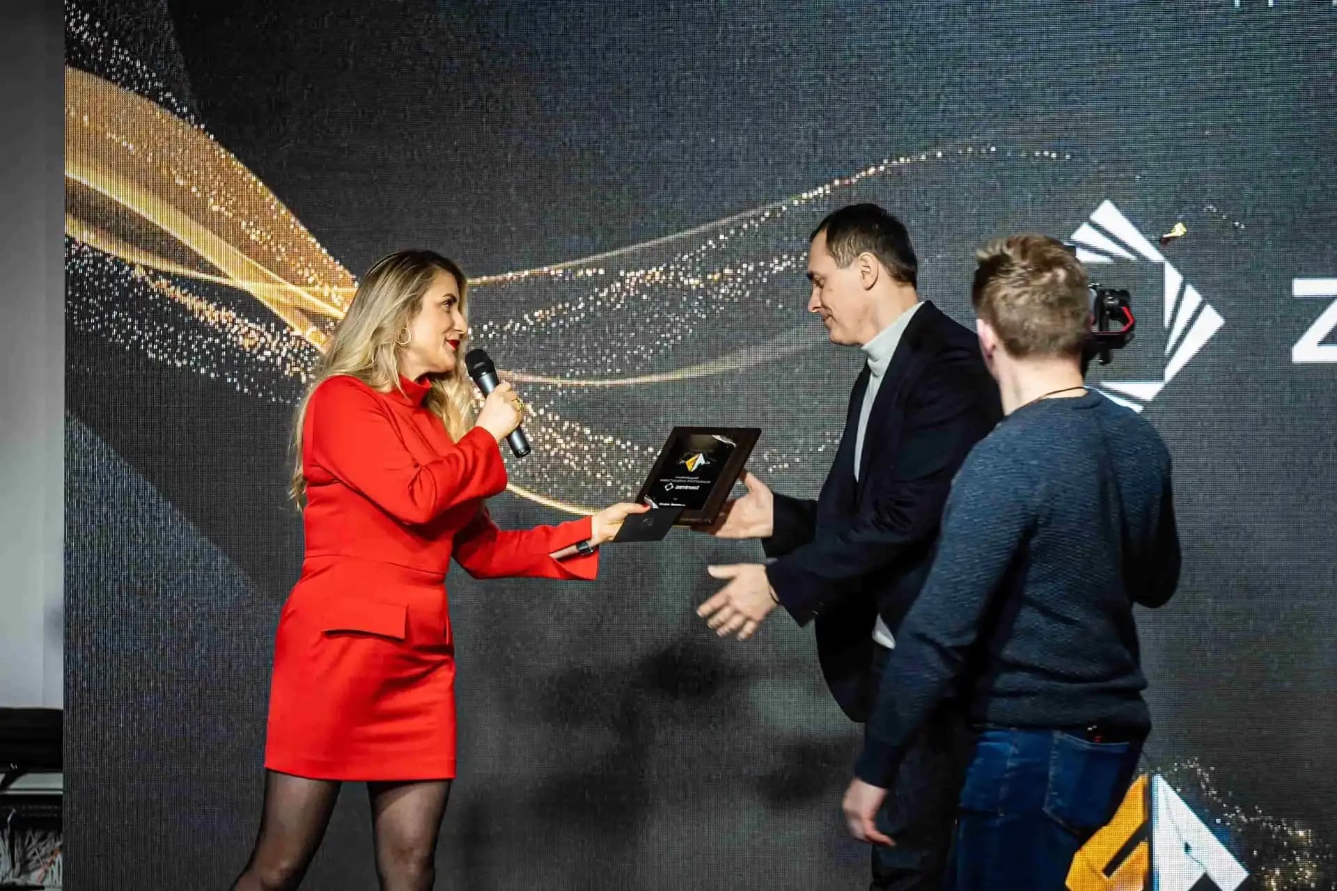 Woman in a red dress presenting an award on stage to a man in a suit, with a cameraman capturing the moment.