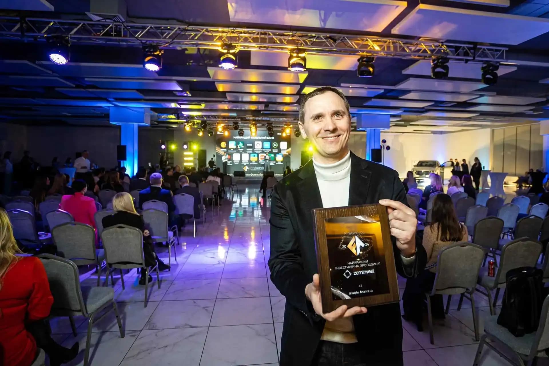 Person with a smile holding an award in a conference room with an audience and stage in the background.