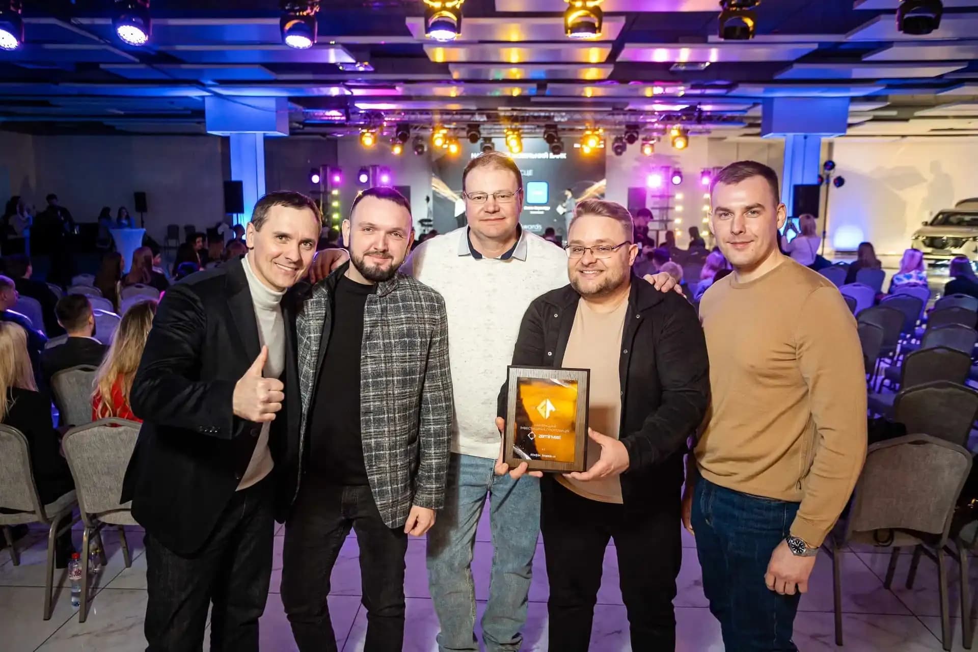 Group of people at an award ceremony holding a plaque, with a vibrant stage and audience in the background.