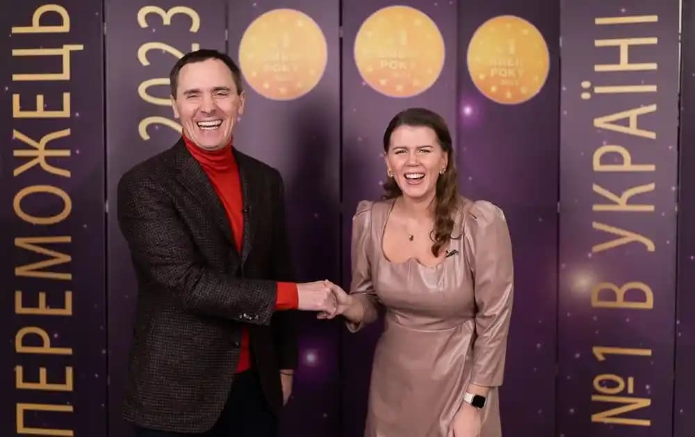 A man and a woman are shaking hands in front of a banner with the words 'Winner 2023' and 'No. 1 in Ukraine'. They are smiling and looking festive.